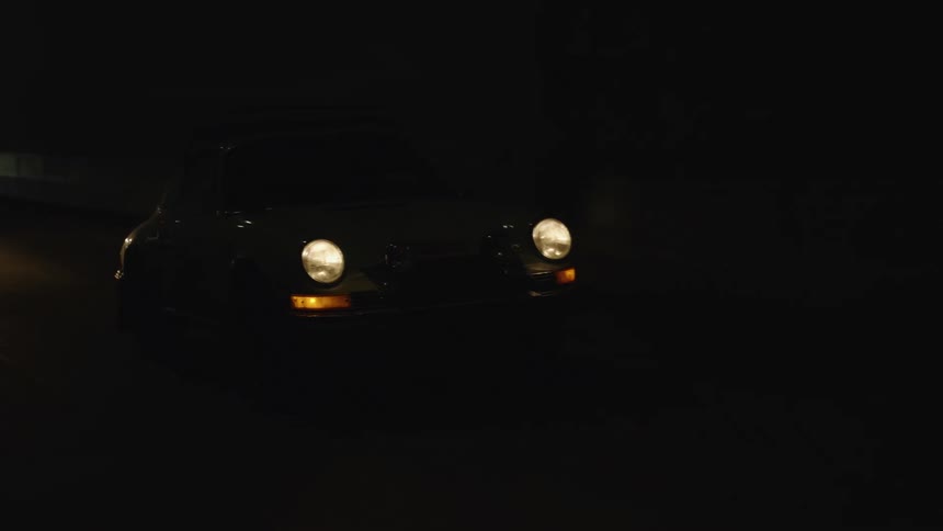 A white vintage racing car with a roof rack and the number seventy nine on the door drives through a tunnel with its headlights on