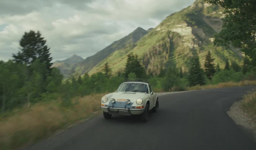 Sports car drives along a highway curving past forests and mountains on a cloudy day