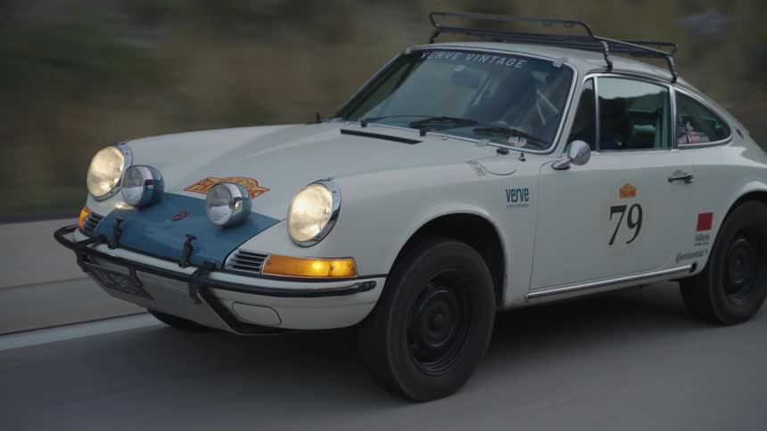 A white vintage racing car with a roof rack and the number seventy nine on the door drives along an asphalt road during a cloudy evening with its headlights on