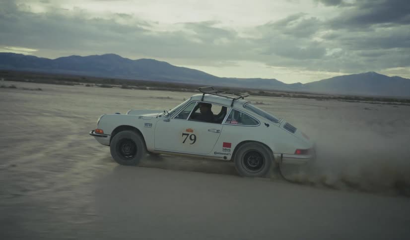 Numbered sports car drives across sandy terrain with mountains in the background