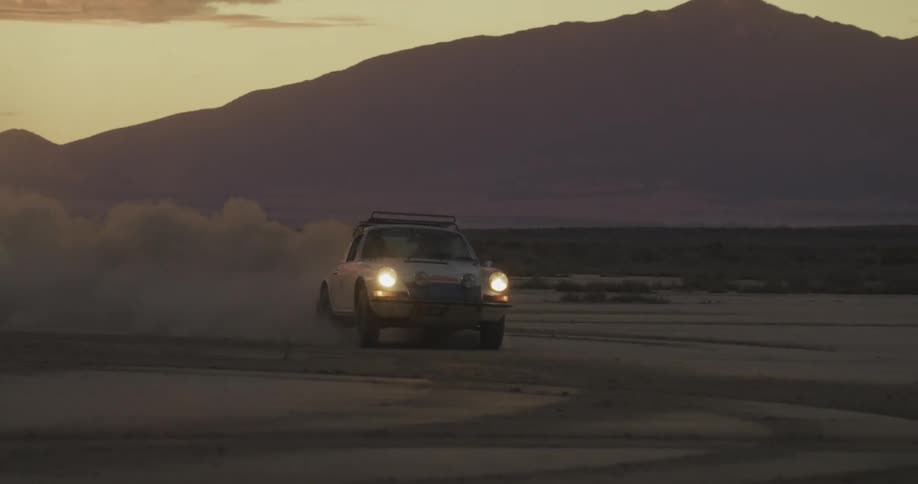 A white vintage rally car drives across a dusty desert plain during sunset and the vehicle kicks up a large cloud of sand as it approaches the camera with its headlights shining against the backdrop of dark purple mountain silhouettes