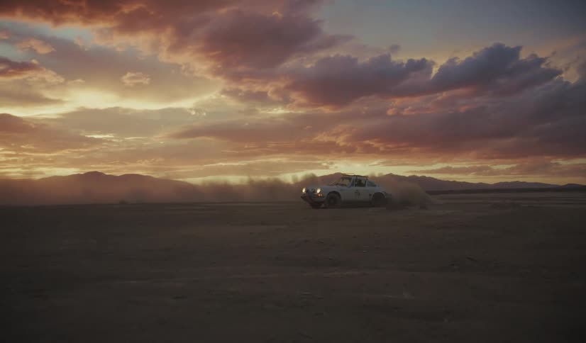 A rally car kicks up dust while driving across a vast desert landscape at dramatic sunset