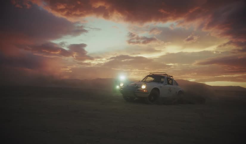 A white vintage racing car and another dark vehicle drive through a flat desert landscape at sunset kicking up thick clouds of dust under a dramatic sky with orange and purple clouds