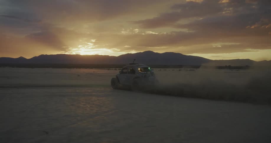 Small car drives across the desert while the sun shines near the silhouetted mountains in the background