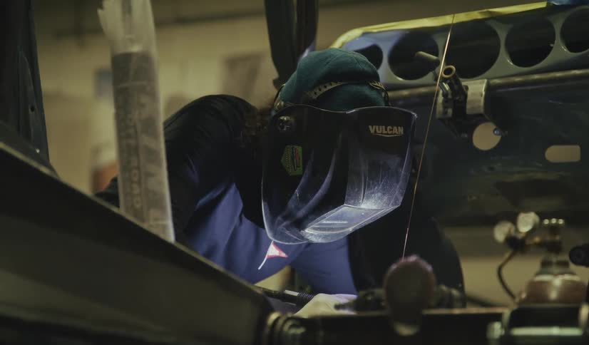 A welder wearing a vulcan welding helmet and gloves leans over a car chassis in a dim garage the bright torch glow illuminating their focused metalwork