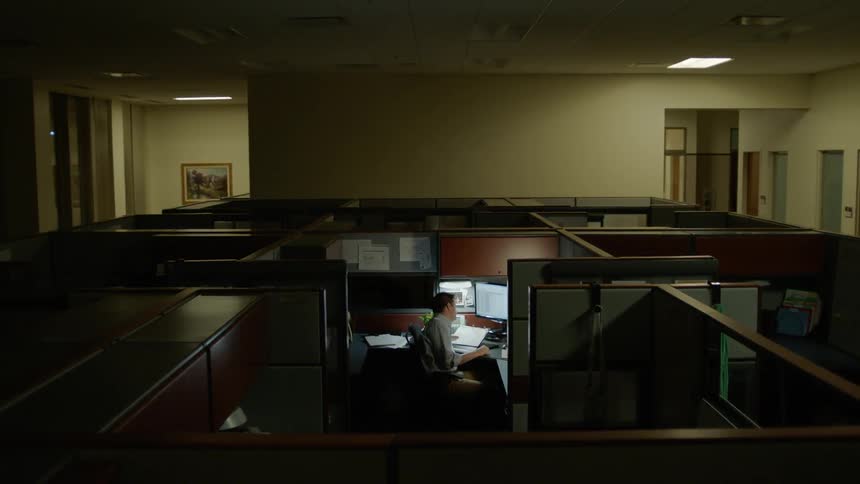 A man works alone in his cubicle as someone turns off the ceiling lights