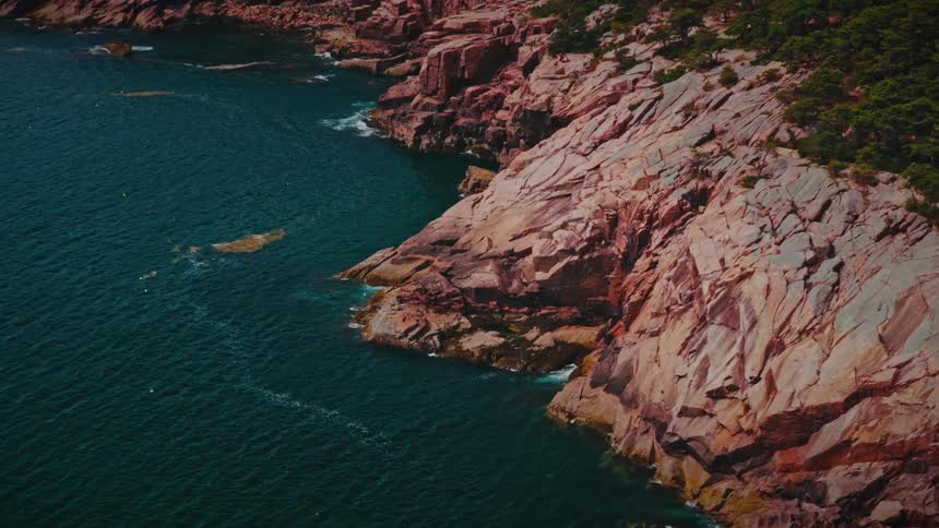 An aerial drone shot slowly tracks right along a rugged coastline where deep blue ocean meets dramatic tree lined rock cliffs