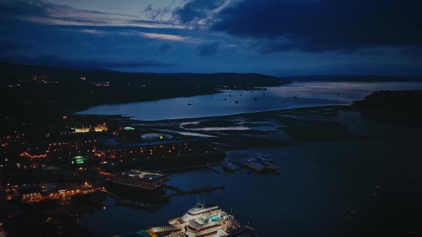 An aerial twilight shot reveals a glowing coastal town nestled beside a calm harbor where several boats are docked