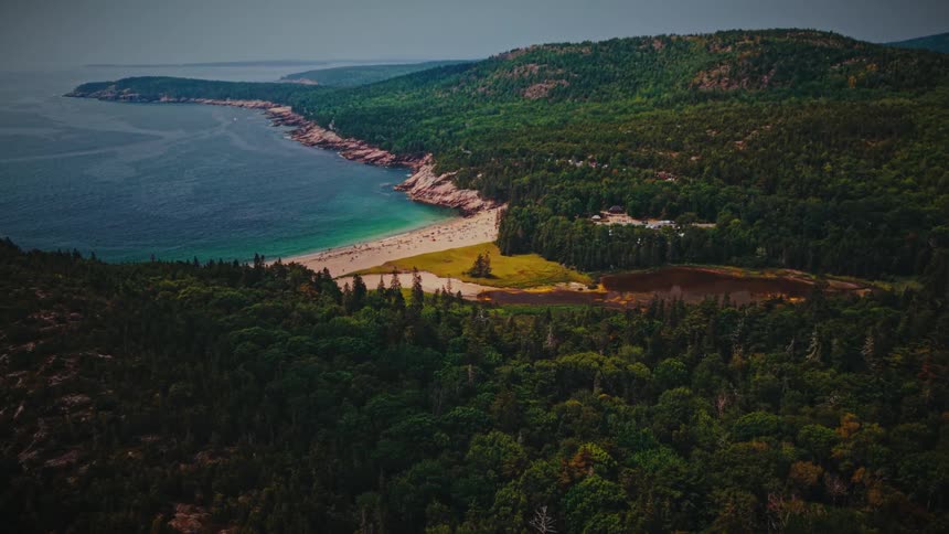 Aerial view of a crescent sandy beach with bright turquoise water tucked between rocky headlands backed by dense forested hills and a small lagoon nestled just inland