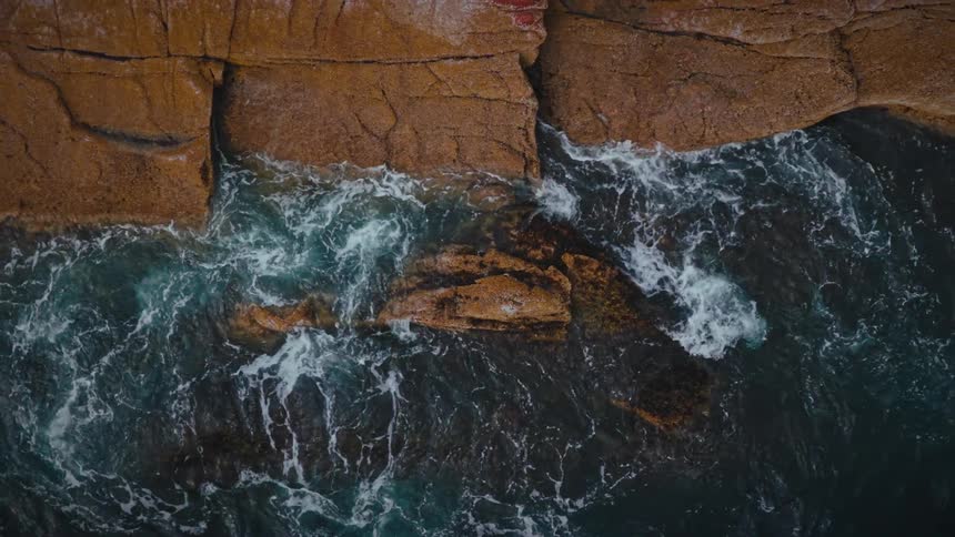 Waves break against orange rock formations seen from above
