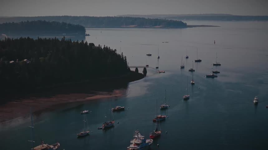 A high angle view shows several sailboats and motorboats anchored in a calm blue bay surrounded by dark green forested islands under a hazy sky during a quiet morning