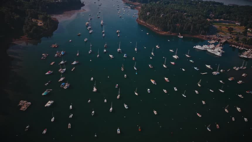 A high angle view shows several sailboats and motorboats anchored in a calm blue bay surrounded by dark green forested islands under a hazy sky during a quiet morning