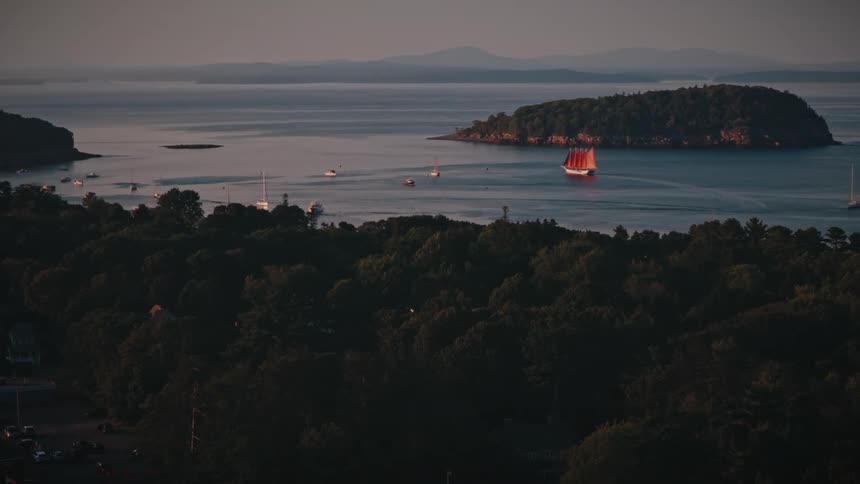 Redsailed schooner sails in a bay with islands and houses among trees in the foreground