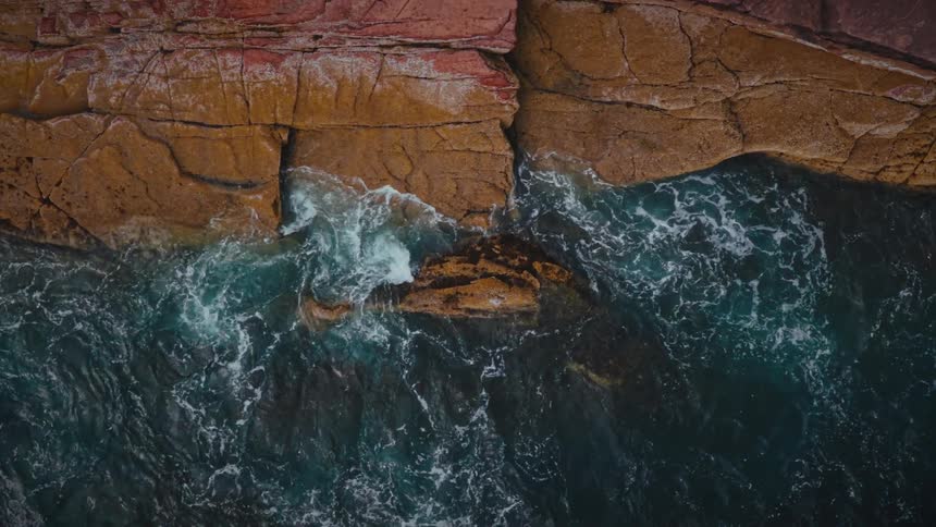 A top view shows dark blue ocean water with white foam crashing against large orange and red rocks along a rugged shoreline