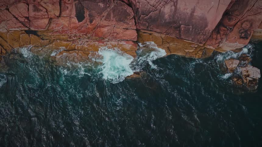 Aerial topdown view of dark teal ocean waves crashing into a jagged reddishbrown rocky shoreline white surf foaming as it pours into cracks and tide pools