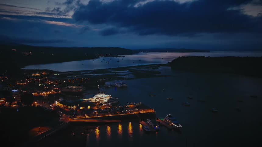 Glowing lights on buildings of a busy coastal community with people and cars on the street and boats sailing on the water
