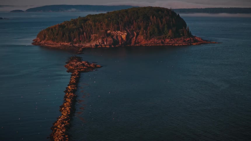 A high angle view shows a small island covered in dense green trees with a long rocky path connecting it to the mainland across deep blue water during a calm sunset