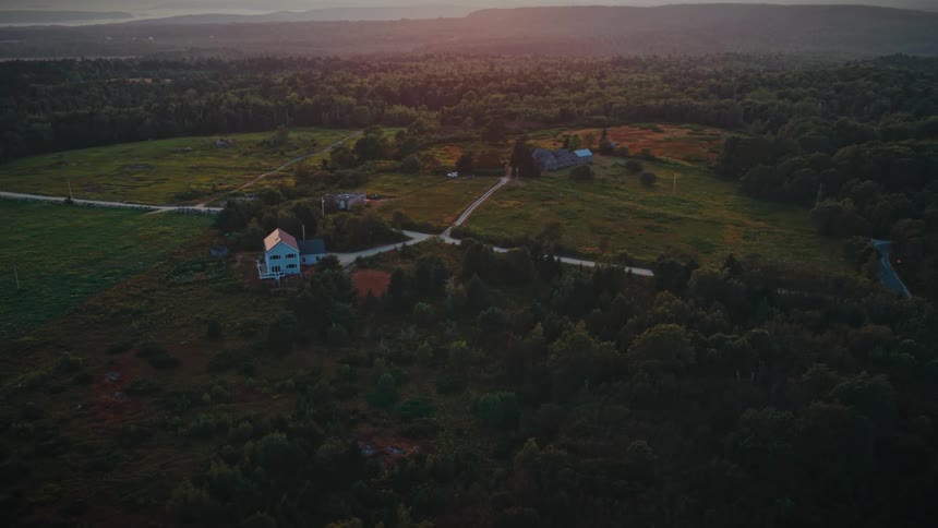 Aerial sunset view of a rural landscape bathed in warm light with a lone house beside a winding dirt road that threads through open fields toward scattered farm buildings and distant forested hills