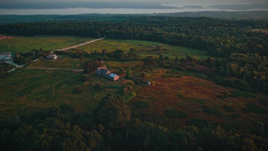 A high angle view shows a large house and several smaller buildings in a grassy clearing surrounded by a dense green forest under a soft sunset sky