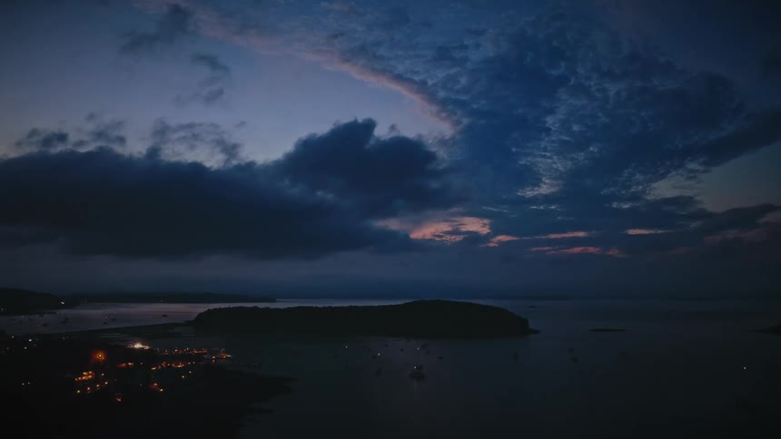 Watercraft sit in the water between settlements in a coastal area and a nearby forested island on a cloudy evening