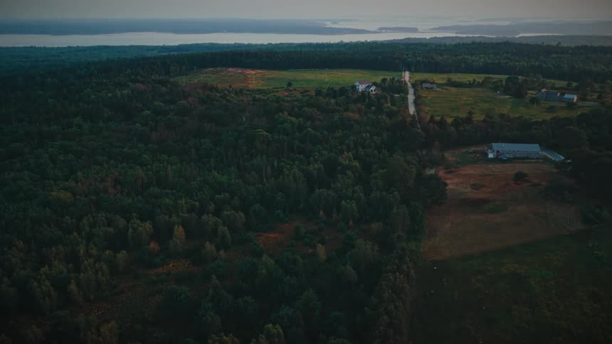 An aerial drone shot slowly pans right over a vast green rural landscape with forests fields and distant water at dawn