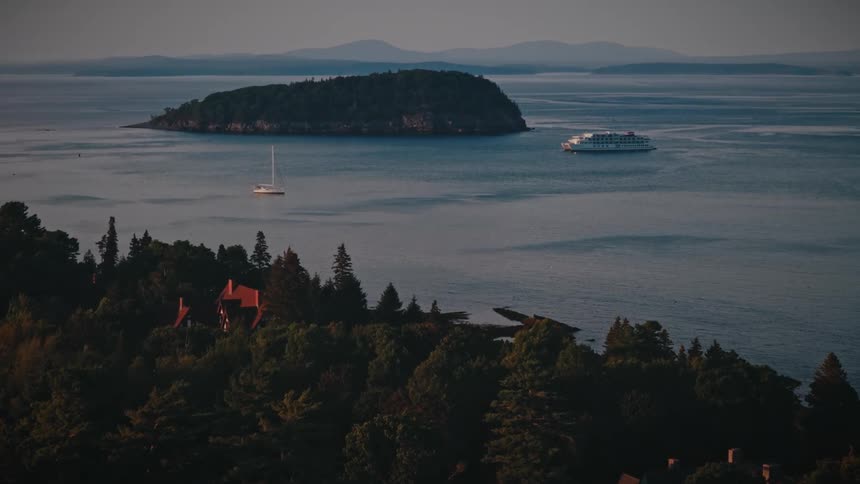 An aerial view shows a large ferry and small sailboats navigating a calm bay near a forested island and a coastline lined with thick green trees and a house with a red roof under a hazy sky with distant mountains in the background