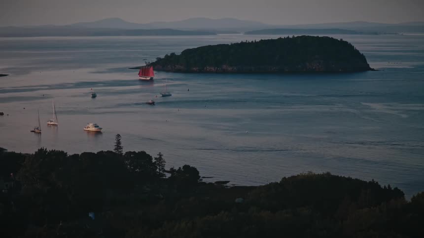 A tranquil coastal dusk scene a treetopped island sits offshore while several boats including a distinctive redsailed vessel bob in calm water with hazy mountains on the distant horizon
