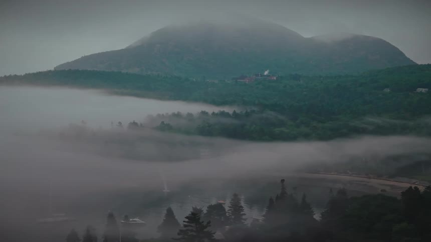 A lake surrounded by green vegetation with a mountain range in the background and a foggy atmosphere on a cloudy day