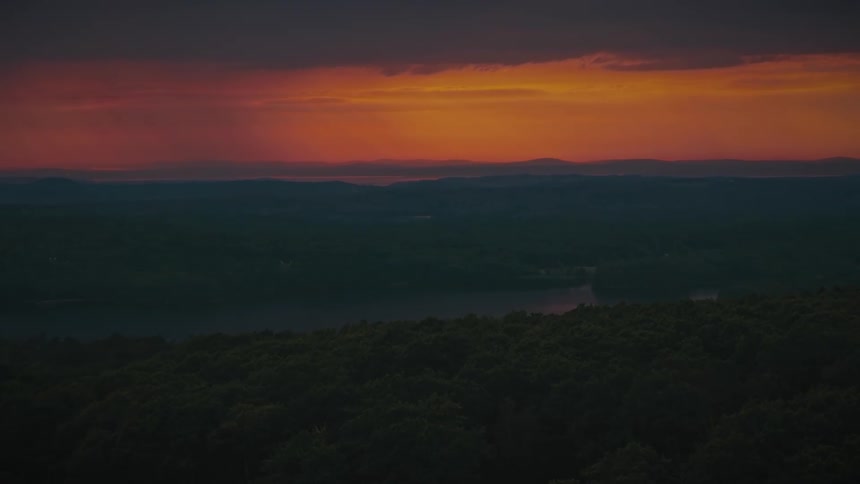 A high angle view shows a small island covered in dense green trees with a long rocky path connecting it to the mainland across deep blue water during a calm sunset