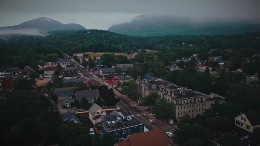 An aerial drone shot slowly pans right over a charming tree filled town nestled beneath cloud shrouded mountains on an overcast day