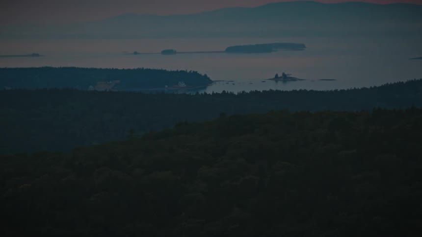A wide high angle view shows a dark green forest in the foreground overlooking a vast body of water at dusk where several small islands and a coastal settlement sit under a hazy purple sky while mountains remain visible in the far distance