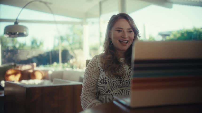 A young woman sitting and having an excited video call on her laptop gesturing with her arms at midday
