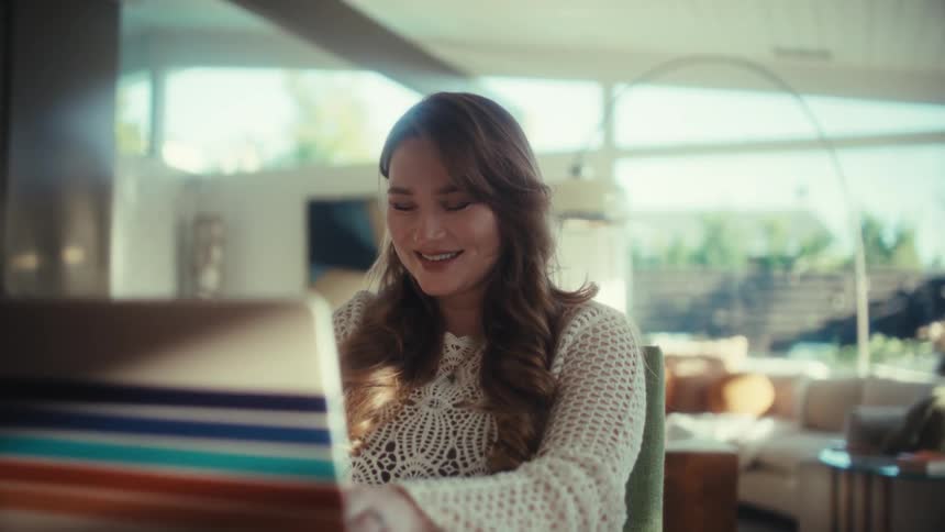 Smiling young adult woman typing on a computer laptop over a wooden table in a living room indoors during day time