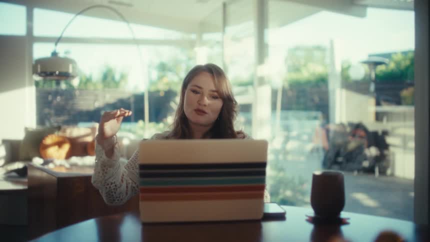 A young woman with long hair sits at a wooden dining table and talks while gesturing with her hands and she is using a laptop in a bright modern room with large windows that look out onto a sunny backyard and patio area