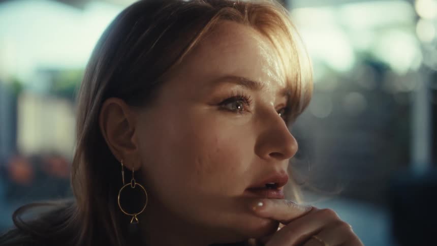 A close view shows a woman with long red hair and gold earrings looking thoughtfully toward the side in a bright indoor setting