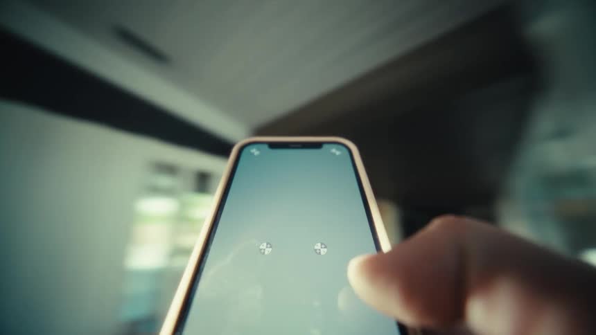 A person holds a smartphone in a bright room while their thumb moves over the screen and sunlight streams through the background where a ceiling fan is visible