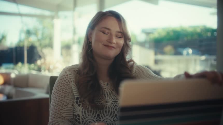 Young adult woman typing on a laptop computer over a wooden table then closing the lid and smiling while sighing in relief indoors during day time