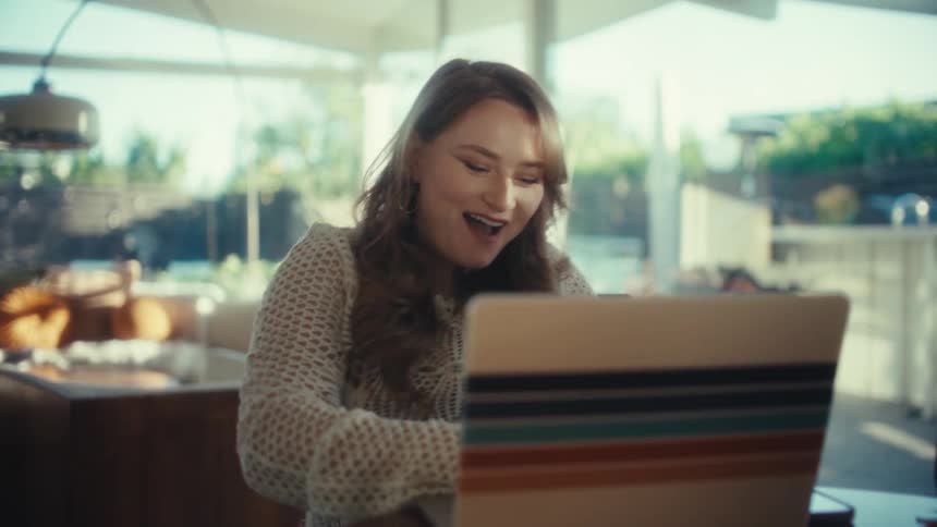 Young adult woman opening the lid of a laptop computer over a wooden table and then smilig and cheering while typing on it indoors during day time
