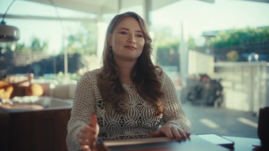 A young woman sits at a wooden table in a sunlit room and closes her colorful laptop before leaning her head on her hand with a tired smile