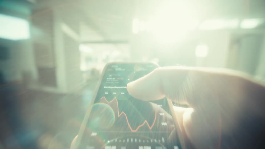 A person walking inside a room at midday holding a smartphone and scrolling through a stock price chart