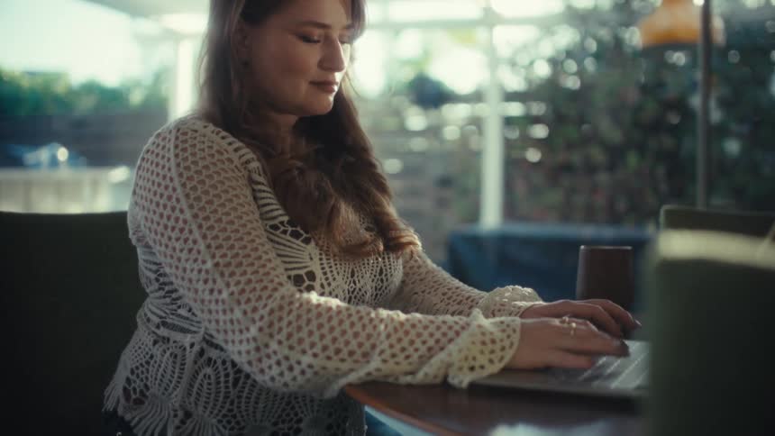 A woman types on a laptop at a table working remotely in a bright modern indoor setting