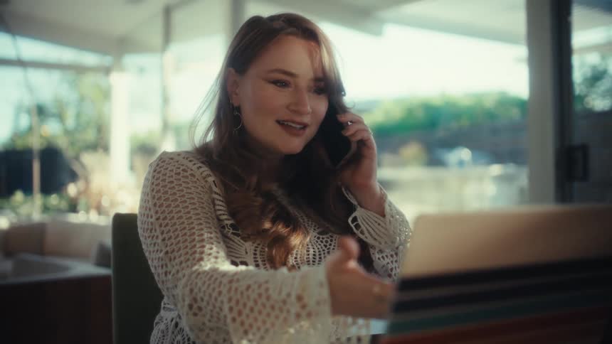 A young woman talks on her smartphone while working indoors on her laptop