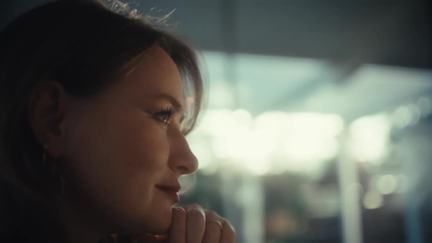 A young woman in a white knit sweater works on her laptop at a desk and she stops typing to rest her chin on her hand looking thoughtfully toward a large window as warm light creates a soft glow around her profile in the modern office
