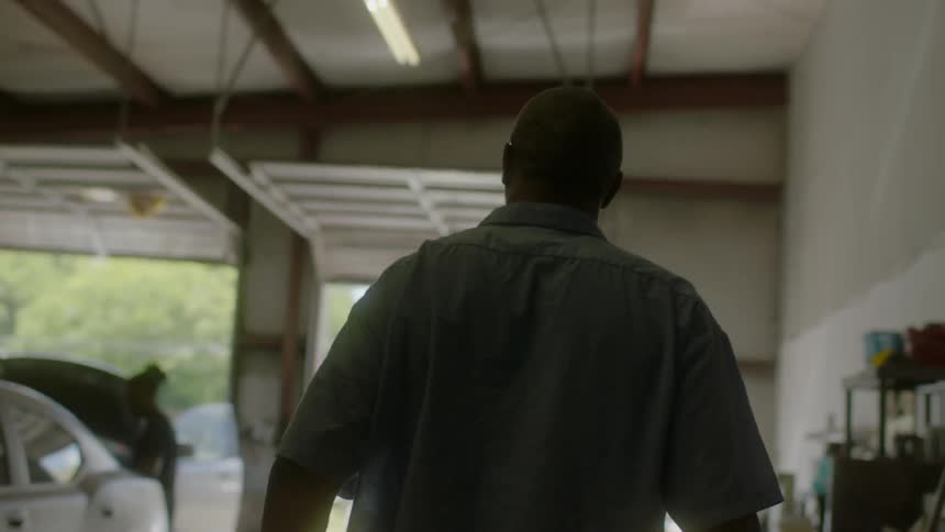 An older man walks into a garage where cars are being worked on