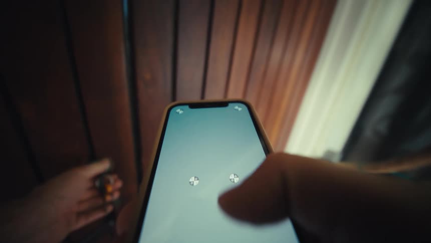 A person holds a smartphone while interacting locking a dark wooden door before stepping outside into a bright daylight area with white walls and green plants