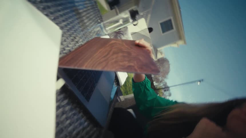 A pregnant woman wearing a green jacket and a pink top sits at a table on an outdoor patio while she closes a laptop with a wooden cover during a bright sunny day with a white house and blue sky visible in the background