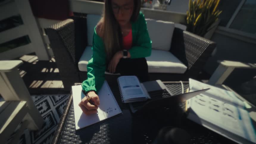 A young woman wearing glasses and a green jacket sits on an outdoor patio writing in a notebook near a laptop and textbooks while working in the bright sunlight