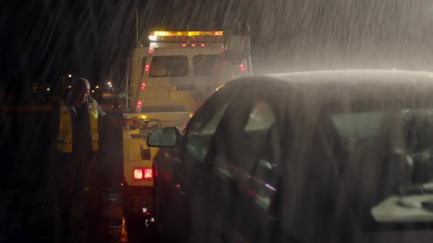 A tow truck driver hooks up a car in heavy rain at night