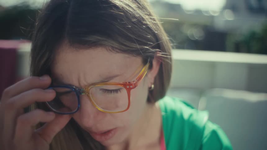 A woman wearing colorful glasses and a green jacket sits outdoors and looks toward the camera with a tired expression before removing her glasses in the bright sunlight