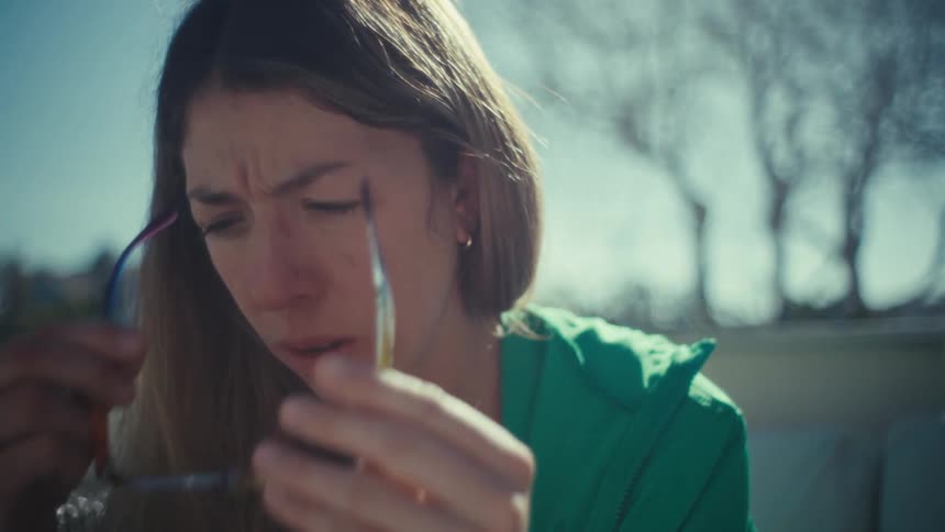 A woman wearing a green jacket and colorful glasses sits outdoors on a patio and rubs her eyes with a tired expression while working in the bright sunlight